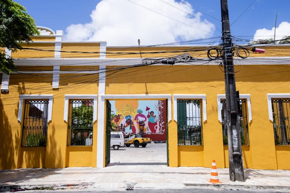 Na imagem, a fachada de um prédio de cor amarelo-ovo forte, compreendendo o Teatro Carlos Câmara. A fachada tem quatro aberturas frontais retangulares com grades de ferro pretas (duas em cada lado) e uma grande abertura central, que parece ser um portão ou passagem, revelando o interior. Os contornos das janelas e portas são brancos e verdes. Através da abertura central, é visível um pátio interno ou área aberta, onde está estacionado um carro amarelo tipo jipe/buggy e uma van branca. Ao fundo, na parede interna, há um grande mural colorido com figuras humanas e folclóricas. No canto direito da foto, há um poste de madeira escuro sobrecarregado de fiação elétrica e cabos, com uma fita isolante rosa no topo e um cone de sinalização laranja e branco na base. O chão é de calçada e há vegetação (árvores) no canto superior esquerdo. O céu está azul com algumas nuvens.