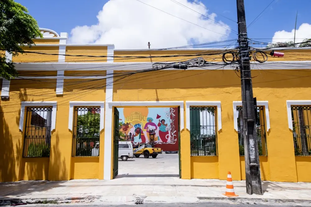 Na imagem, a fachada de um prédio de cor amarelo-ovo forte, compreendendo o Teatro Carlos Câmara. A fachada tem quatro aberturas frontais retangulares com grades de ferro pretas (duas em cada lado) e uma grande abertura central, que parece ser um portão ou passagem, revelando o interior. Os contornos das janelas e portas são brancos e verdes. Através da abertura central, é visível um pátio interno ou área aberta, onde está estacionado um carro amarelo tipo jipe/buggy e uma van branca. Ao fundo, na parede interna, há um grande mural colorido com figuras humanas e folclóricas. No canto direito da foto, há um poste de madeira escuro sobrecarregado de fiação elétrica e cabos, com uma fita isolante rosa no topo e um cone de sinalização laranja e branco na base. O chão é de calçada e há vegetação (árvores) no canto superior esquerdo. O céu está azul com algumas nuvens.