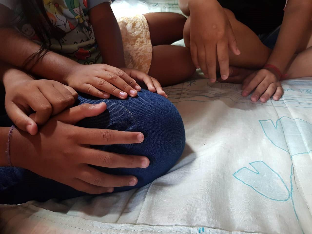 Foto mostra detalhes de mãos de três pessoas sentadas num colchão. Uma delas é uma mulher adolescente e outras duas são de crianças, provavelmente filhos dela, revelando uma situação de união conjugal precoce no Ceará.