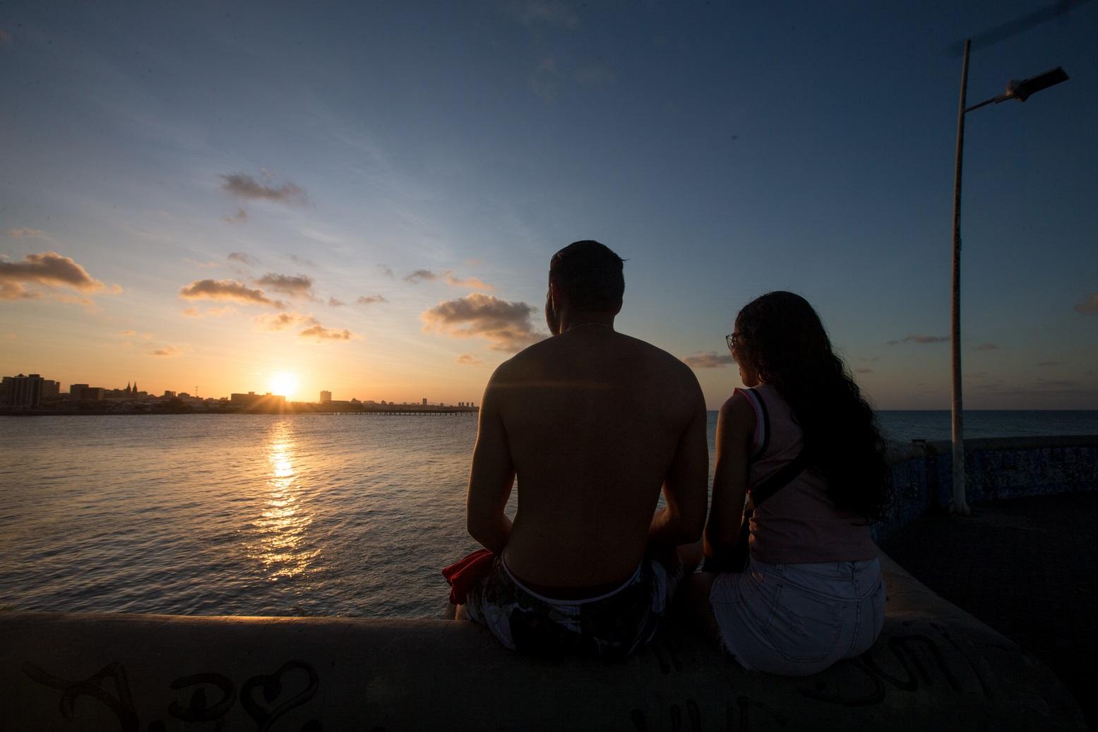 Foto de silhueta de casal sentado na parede de um espigão de Fortaleza em frente ao mar, ao pôr do sol. O homem, à esquerda, usa bermuda e está sem camisa. A mulher usa camiseta e óculos. Ao fundo, é possível ver detalhes de prédios da orla.