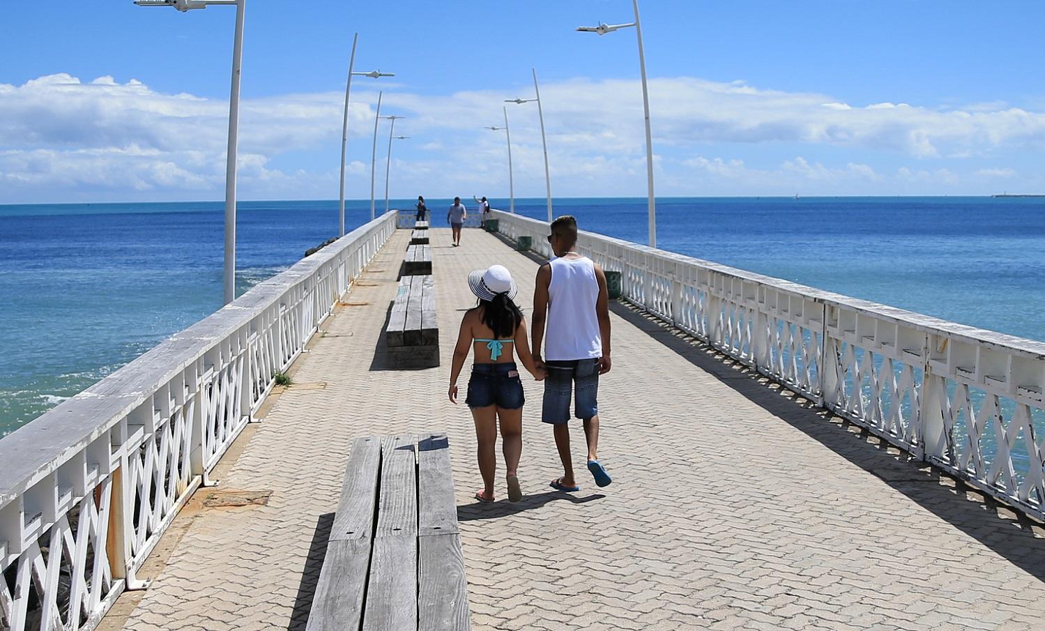 Foto de um casal heterossexual caminhando de mãos dadas em um espigão na Beira-Mar, sob um céu azul e ensolarado. O mar rodeia a passarela branca com bancos ao centro. Algumas pessoas aparecem ao fundo, aproveitando o dia à beira do oceano Atlântico.