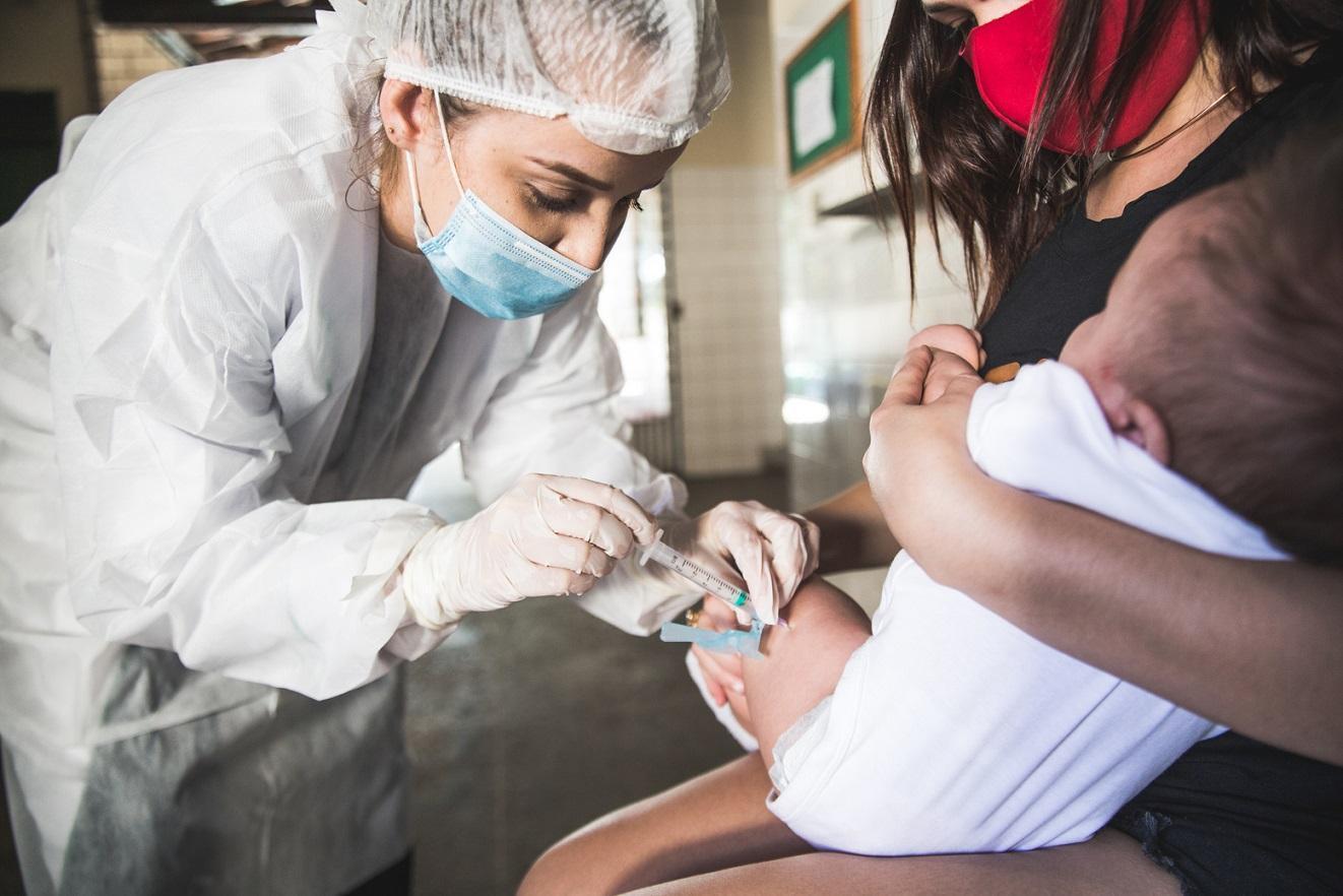 Close-up de uma profissional de saúde vacinando um bebê no braço. A criança está no colo de uma mulher. A imagem foca no momento da injeção, sugerindo vacinação infantil, destacando o cuidado e a segurança.