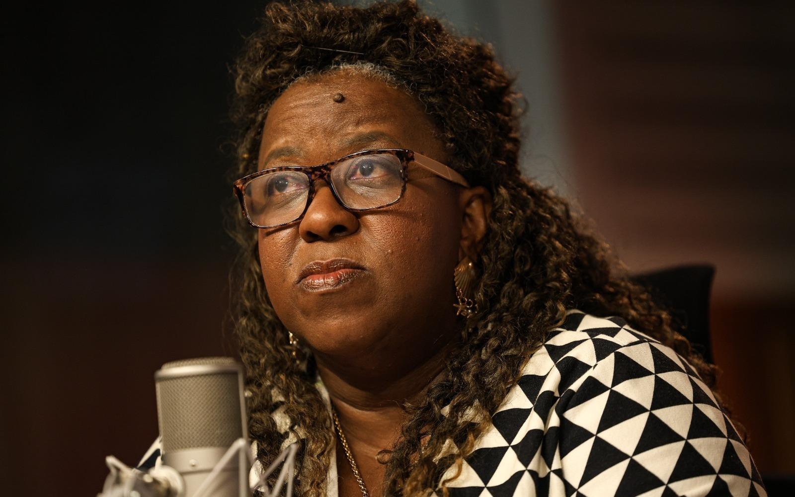 Macaé Evaristo é uma mulher negra, de meia idade, que usa óculos retangulares de acetato. Na foto, ela fala ao microfone e está com o semblante sério. Usa blusa de formas geométricas em branco e preto.