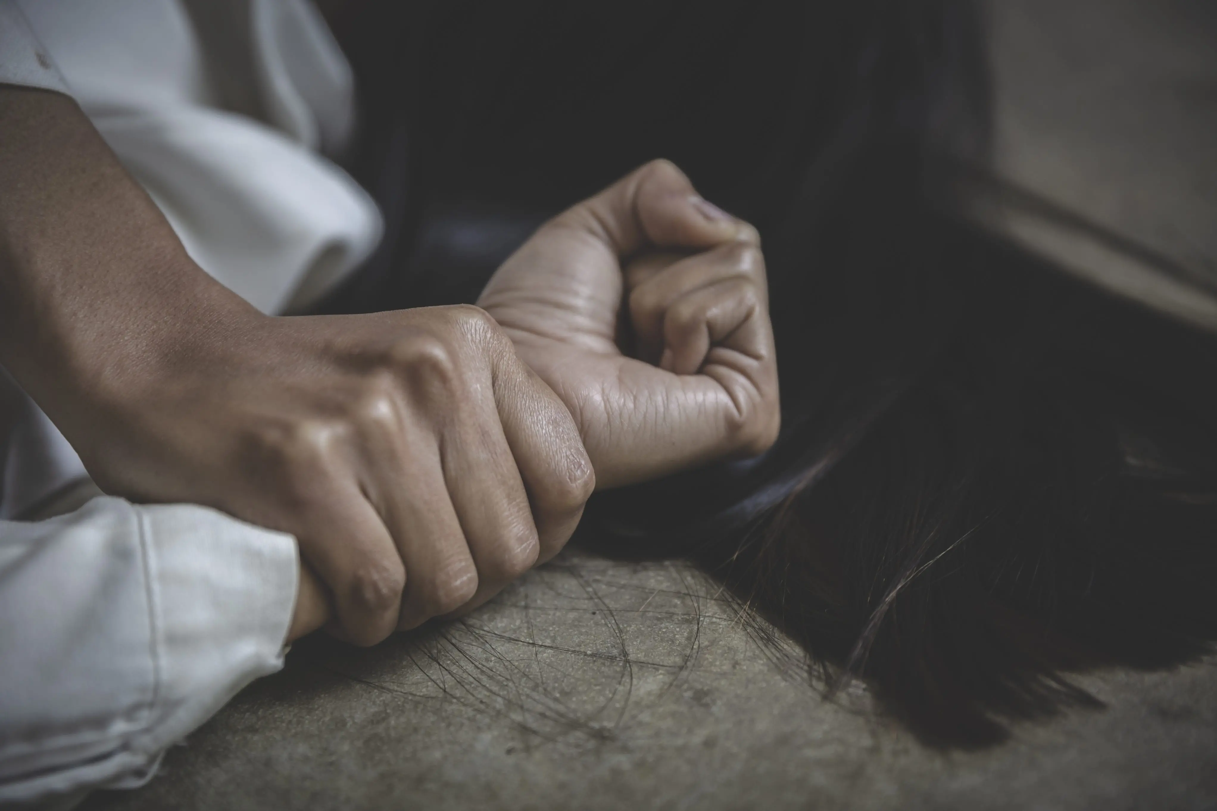 Close-up de uma pessoa com a mão sendo segurada com força por outra, sugerindo uma situação de violência ou agressão. A cena é tensa e mostra apenas as mãos e parte do cabelo da vítima, deitada no chão.