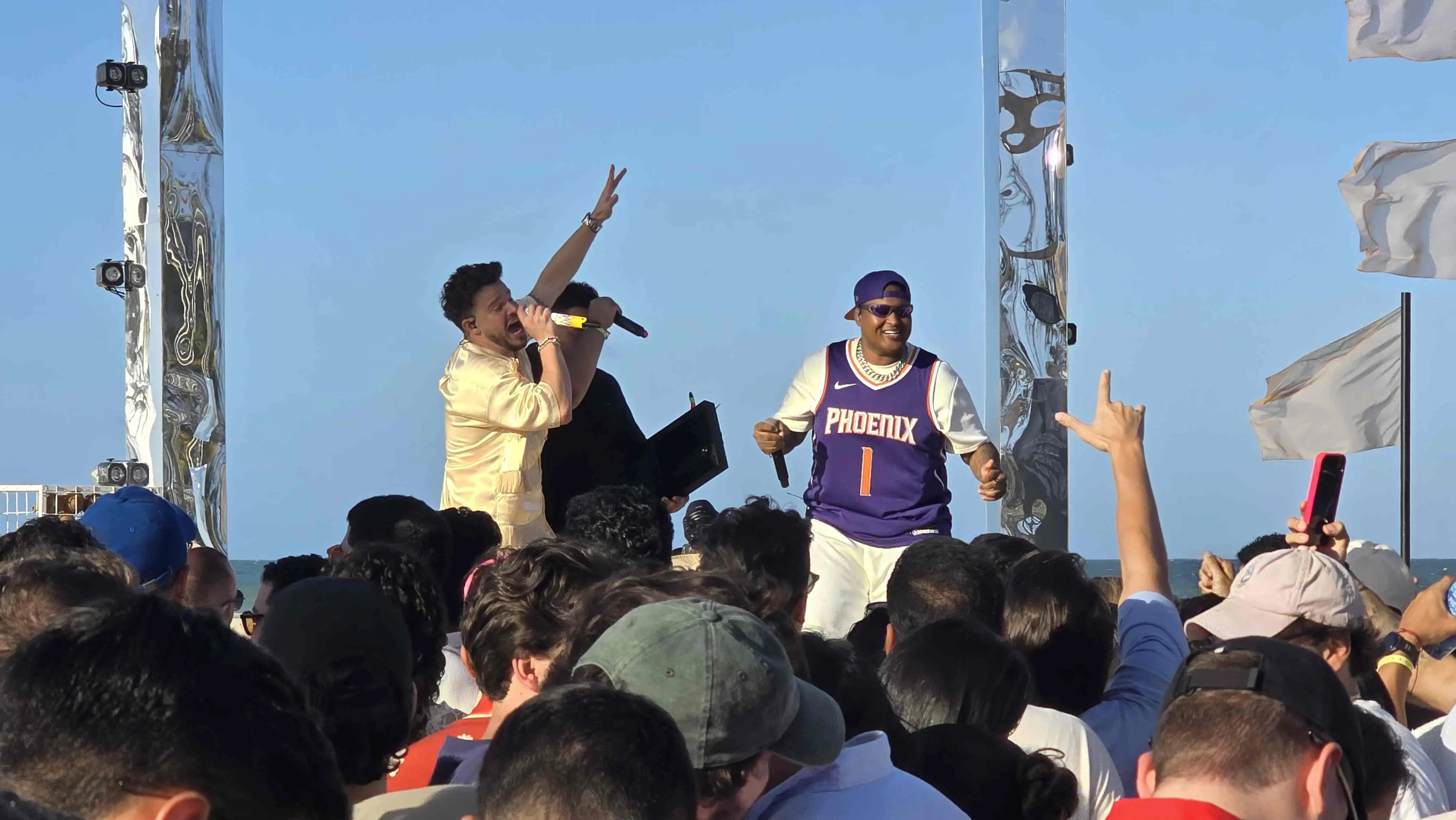 Kevin o Chris ao lado de Matheus Fernandes no palco do DVD 'Fala Meu Nome'. O fundo da imagem é um céu azul. Na beira do palco, o público ergue as mãos acompanhando a música.