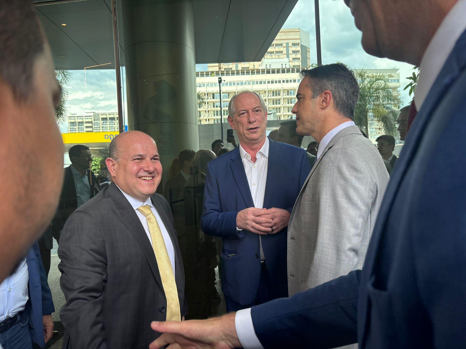 Roberto Cláudio, Ciro Gomes e Capitão Wagner em trajes formais conversam sorrindo na entrada de um prédio envidraçado. A cena mostra um ambiente de evento político ou institucional, com outros participantes ao fundo e um dia ensolarado refletido nas janelas.