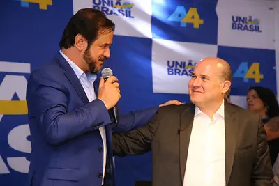 Antônio Rueda está em pé ao lado de Roberto Cláudio. O dirigente partidário está em um salão cheio de espectadores durante evento de filiação do ex-prefeito de Fortaleza ao partido. Rueda veste uma camisa azul clara com um blazer azul escuro por cima, segura um microfone e olha para Roberto Cláudio.