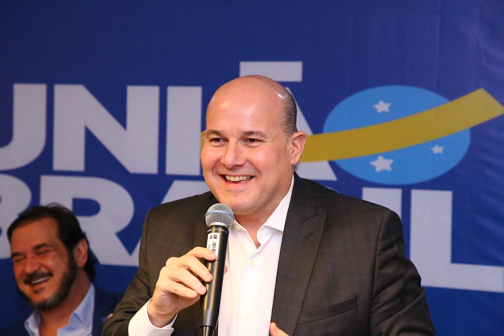 Roberto Cláudio, político brasileiro, em um evento do partido União Brasil. Ele está em primeiro plano, usando um terno escuro sobre uma camisa social branca, segurando um microfone com a mão direita e gesticulando com a esquerda. Ao fundo, há um painel azul e branco com a logomarca do partido 