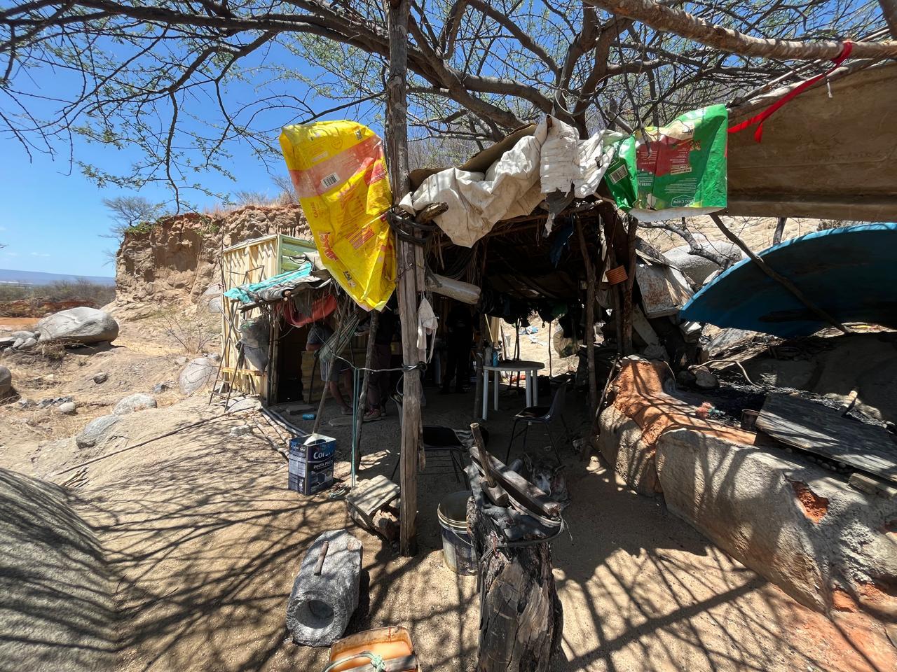 Barraco de lona sustentado por galhos onde morava o idoso resgatado. Na foto, é possível ver ainda sacolas plásticas penduradas sob o sol.