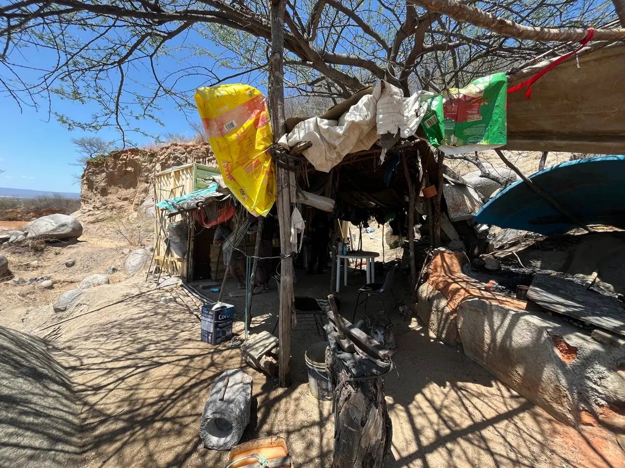Barraco de lona sustentado por galhos onde morava o idoso resgatado. Na foto, é possível ver ainda sacolas plásticas penduradas sob o sol.