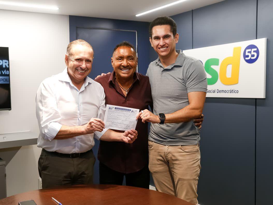 Homens em um escritório posam sorridentes para a foto. O homem ao centro segura uma ficha de filiação partidária, com os outros dois segurando as extremidades do documento. Atrás deles há uma parede azul com o logotipo do partido PSD (Partido Social Democrático) e o número 55. A cena transmite um momento de celebração, possivelmente referente a uma nova filiação política.