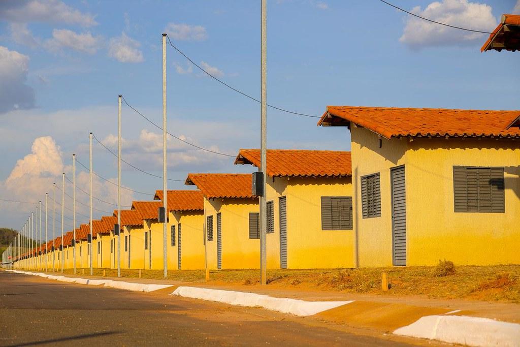 Imagem de casas residenciais com telhados vermelhos, paredes amarelas, janelas com persianas cinzas e postes de iluminação ao longo da rua em uma cidade tranquila.