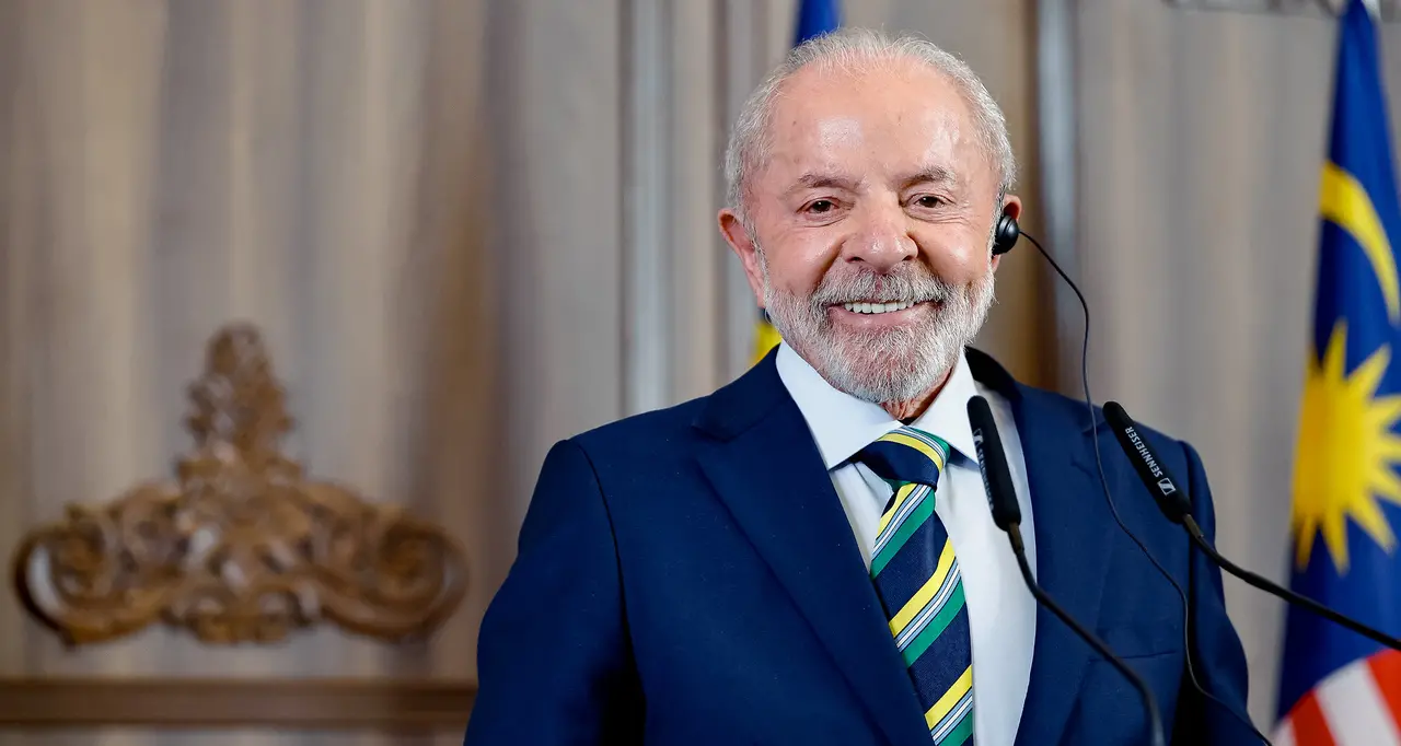 Imagem mostra Luiz Inácio Lula da Silva, presidente do Brasil, sorrindo em um evento oficial, vestindo terno azul e gravata com as cores do Brasil, com microfones à frente.