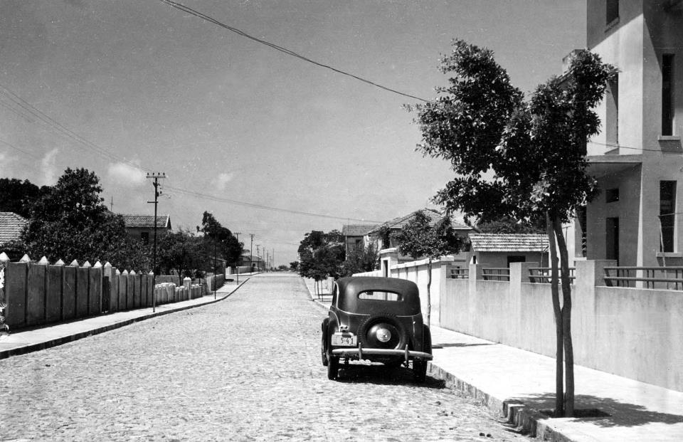 Imagem dos anos 1930 mostra rua Gonçalves Ledo, em Fortaleza. Rua é de calçamento, com casas de muro baixo. À direita, um carro antigo aparece na imagem.