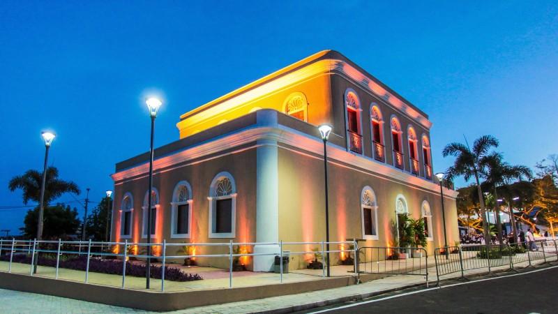 Registro do Memorial Edson Queiroz em Cascavel, Ceará, um prédio de dois andares, com fachada ocre e arquitetura clássica, que aparece está artisticamente iluminado por luzes amarelas sob um céu azul escuro. Palmeiras e postes de luz cercam a estrutura.