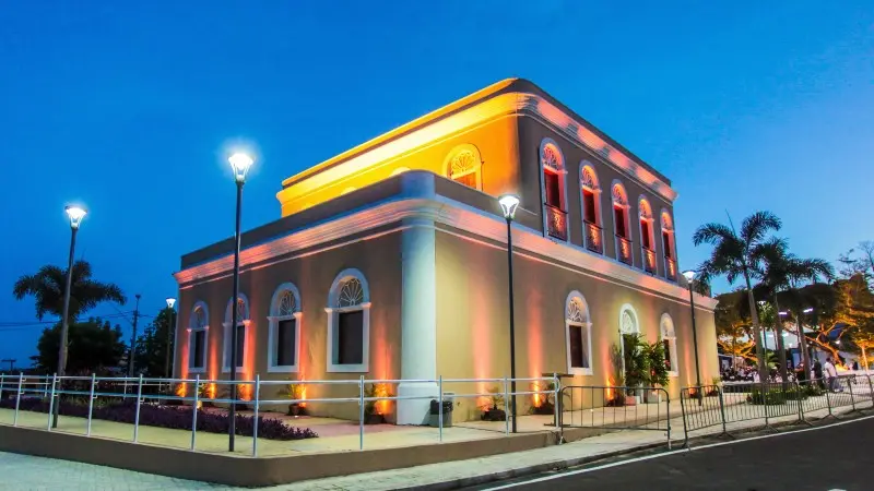 Registro do Memorial Edson Queiroz em Cascavel, Ceará, um prédio de dois andares, com fachada ocre e arquitetura clássica, que aparece está artisticamente iluminado por luzes amarelas sob um céu azul escuro. Palmeiras e postes de luz cercam a estrutura.