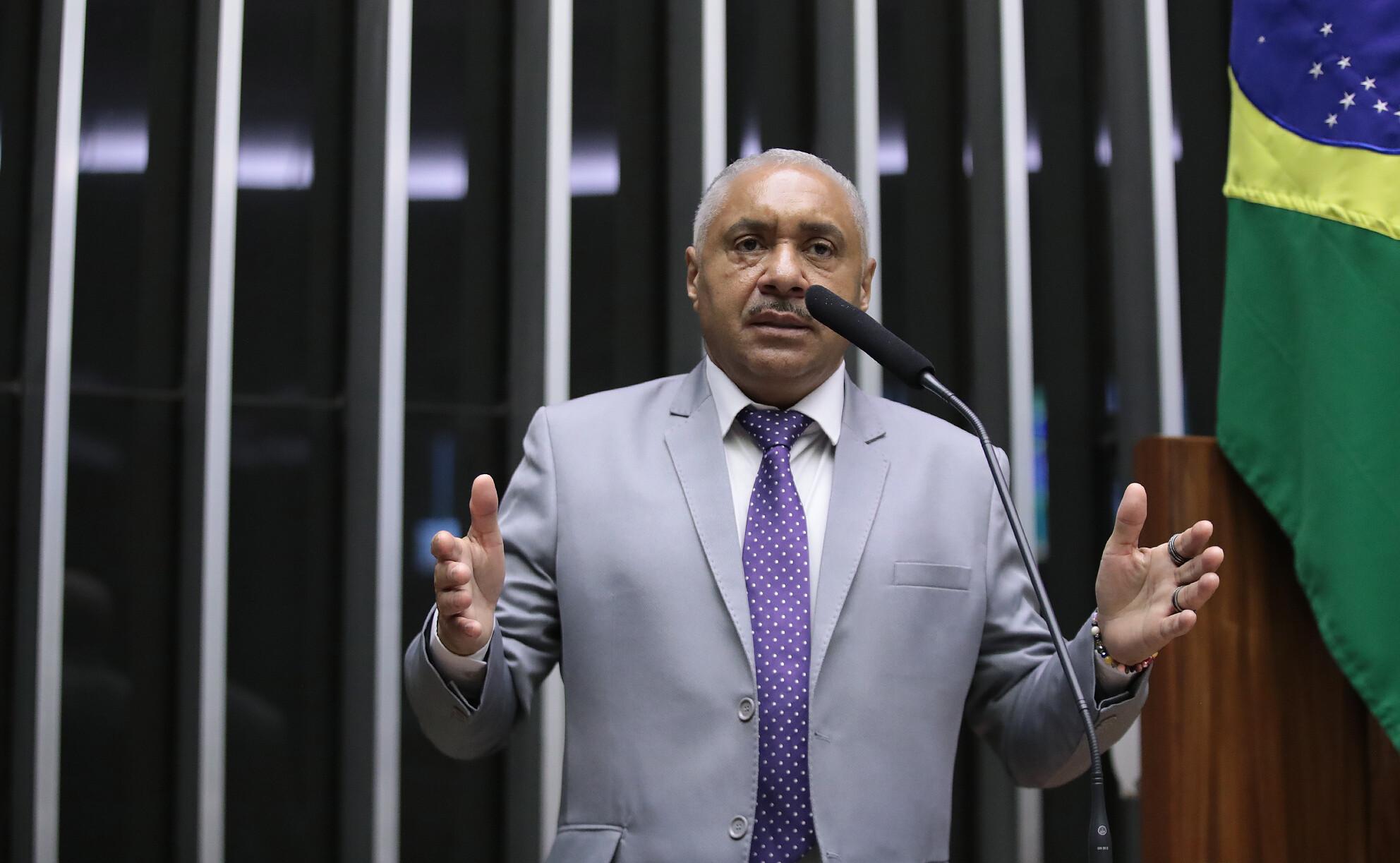 Homem de terno cinza e gravata roxa com bolinhas brancas discursa em um púlpito, gesticulando com as mãos abertas. Ao fundo, há listras verticais escuras e parte da bandeira do Brasil visível à direita da imagem.