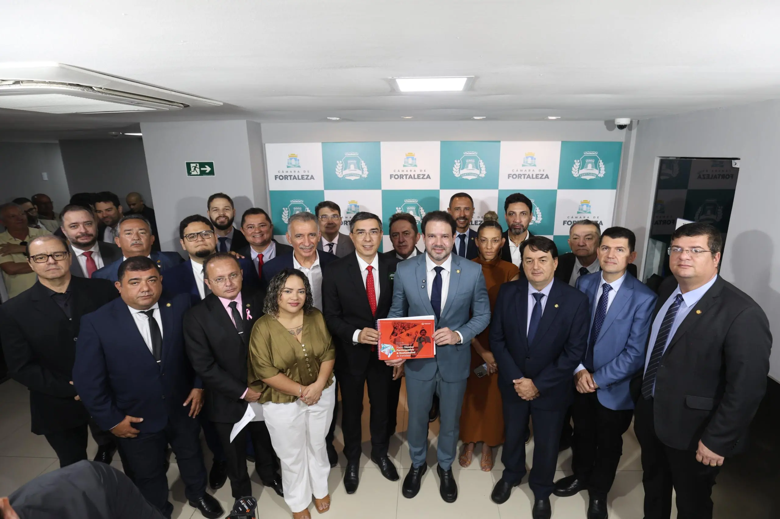 Grupo de cerca de vinte pessoas posa para foto em um ambiente interno, diante de um painel com o logotipo da Câmara Municipal de Fortaleza. No centro, dois homens em trajes formais seguram um caderno de capa vermelha com o título parcialmente visível. As pessoas ao redor, entre homens e mulheres, estão vestidas com roupas sociais e sorriem para a câmera, indicando um evento oficial ou institucional.
