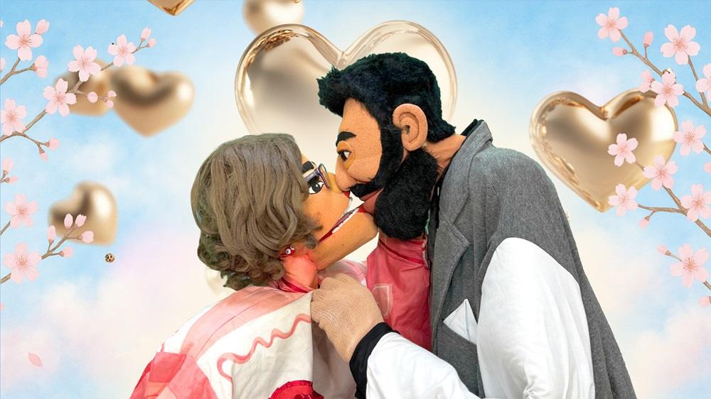 Na imagem, dois bonecos, um com cabelo grisalho e óculos e o outro com barba preta e casaco cinza, se beijando. Eles estão em frente a um fundo azul e branco com corações dourados flutuantes e galhos de flores de cerejeira cor-de-rosa.