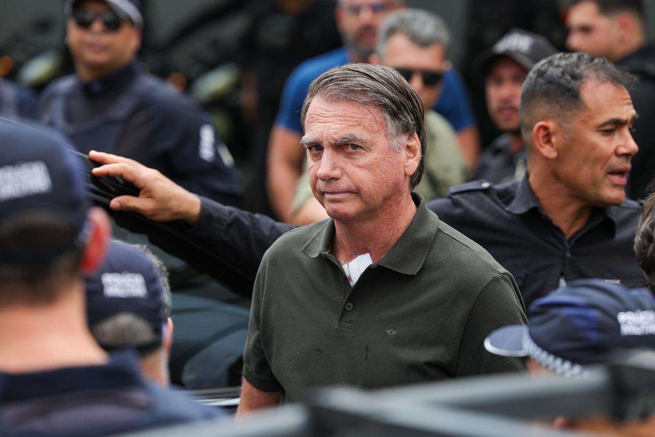 Ex-presidente Jair Bolsonaro com expressão séria, usando camisa polo verde e um curativo visível no pescoço, cercado por agentes de segurança.