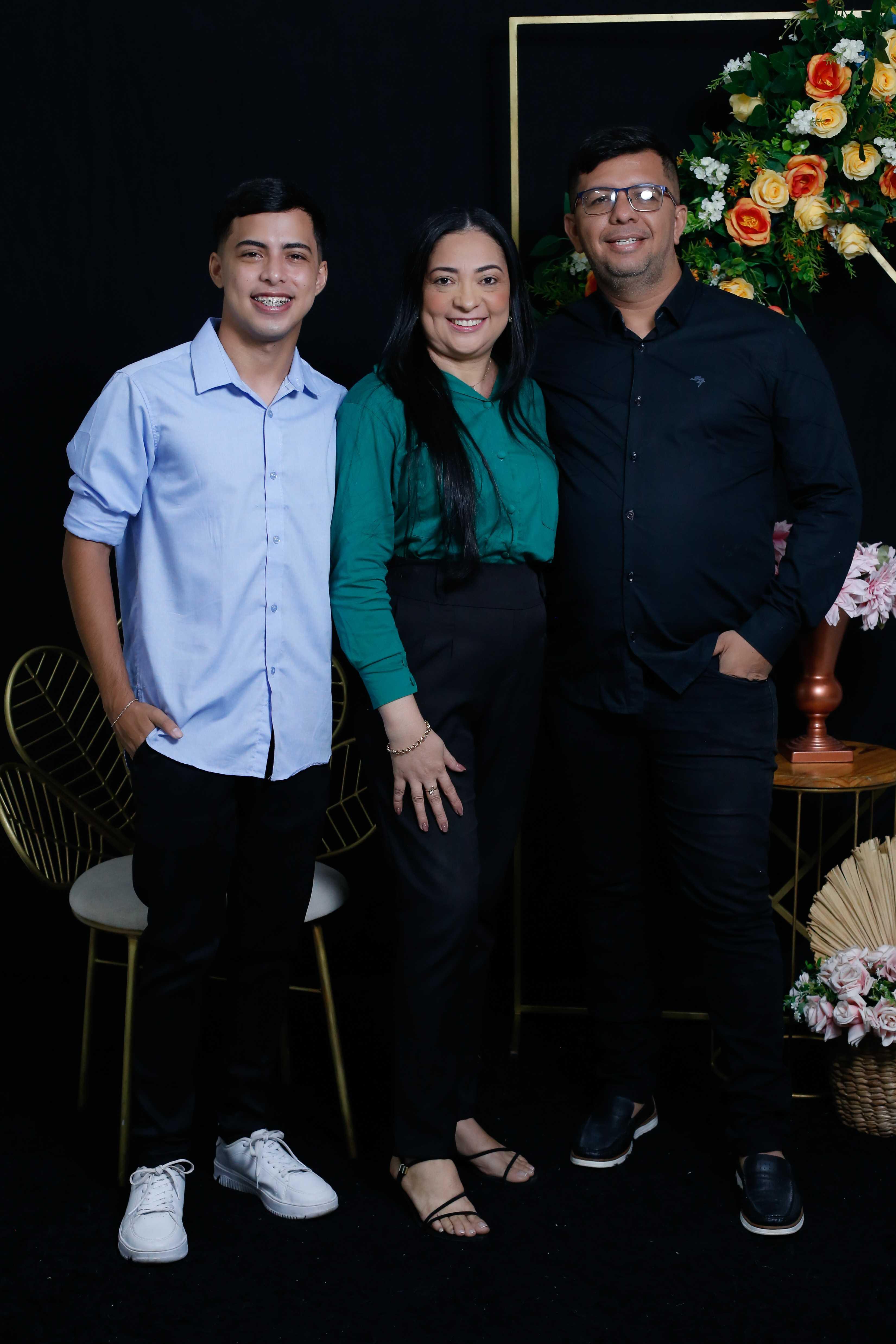 Imagem mostra três pessoas posando: o filho, um jovem com camisa social azul; a mãe, uma senhora de cabelos pretos longos e camisa verde; e o pai, um senhor alto de camisa social preta. Os três sorriem.