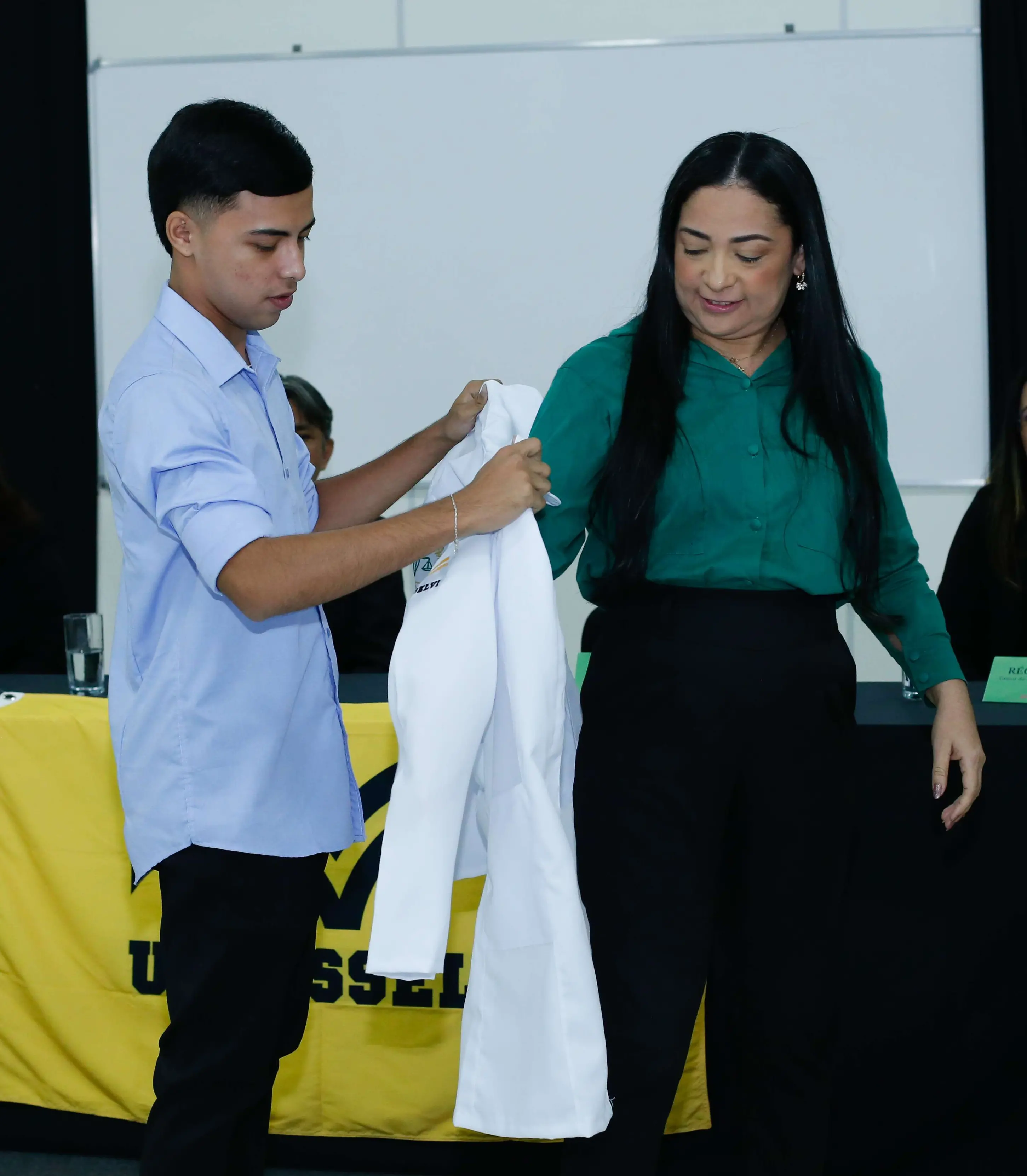 Imagem mostra filho colocando jaleco na mãe durante cerimônia em universidade. Ele é um jovem e veste calça preta e camisa social azul. A mulher tem cabelos pretos longos e veste calça preta e camisa verde.