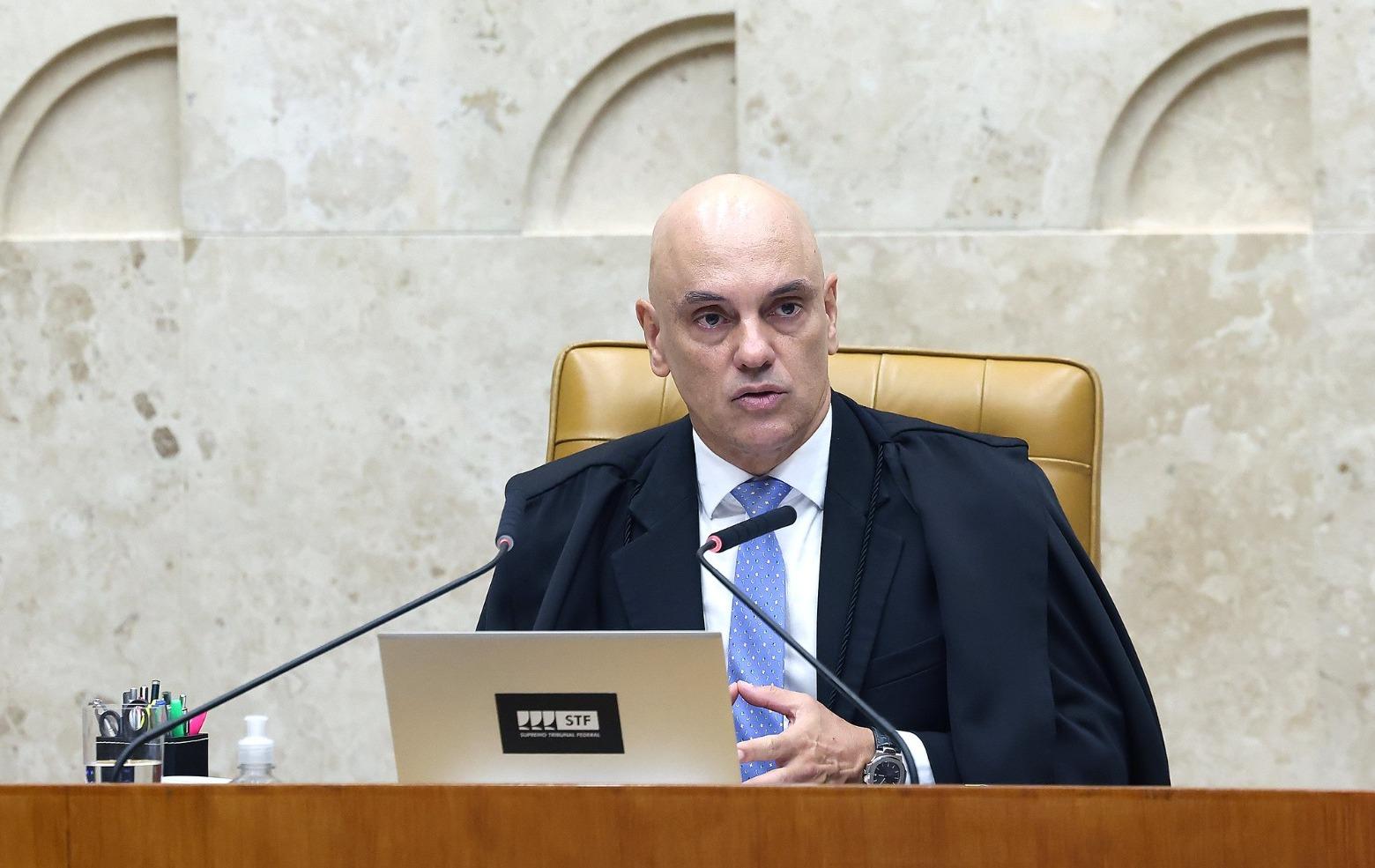 Alexandre de Moraes é um homem de meia idade, branco, careca e, na foto, usa terno preto, camisa social branca e gravata azul. Ele está discursando no plenário do STF.