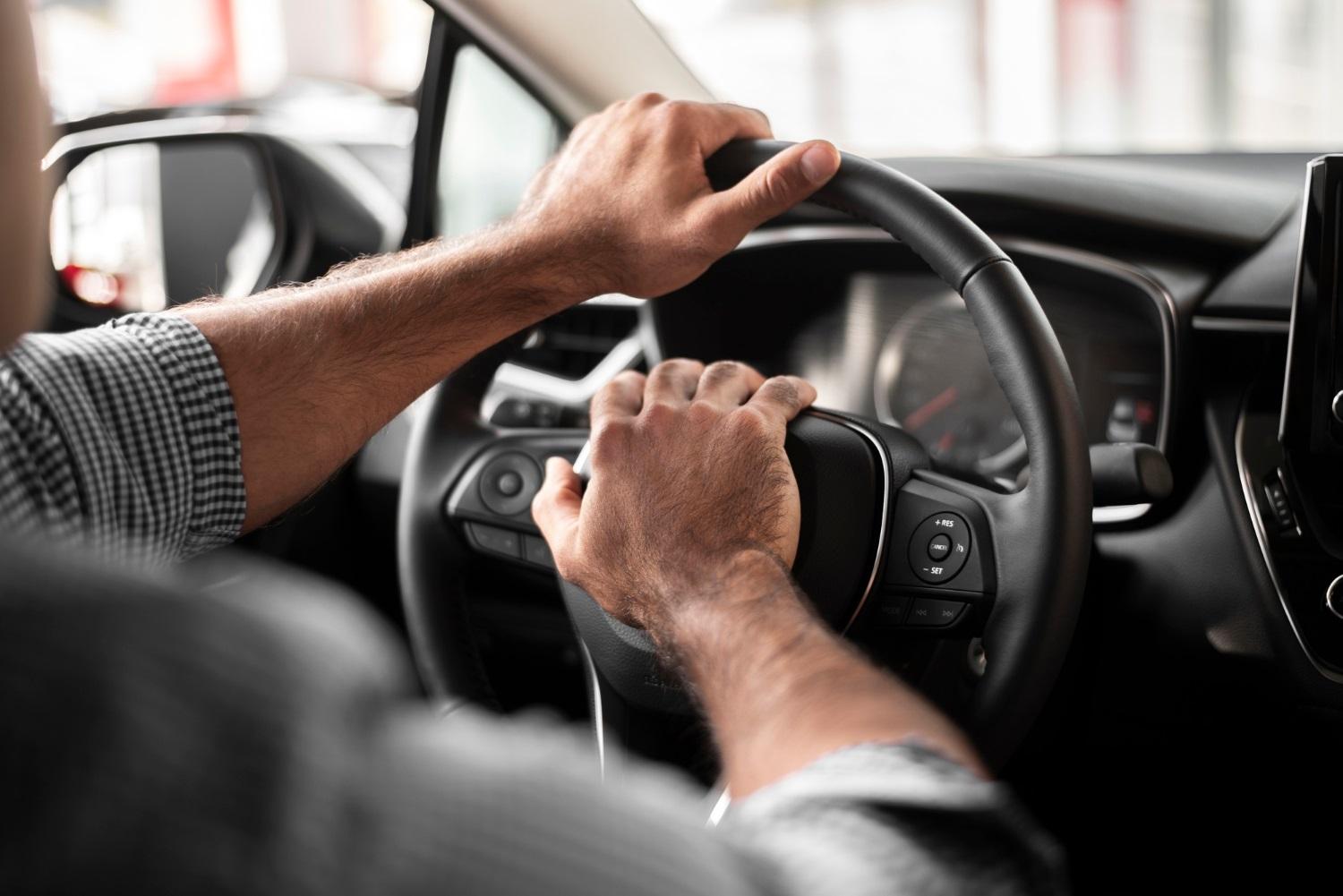 Foto aproximada de mãos de homem segurando um volante e pressionando a buzina, durante um dia ensolarado. O painel do carro é preto e o condutor usa uma camisa social quadriculada e dobrada até o cotovelo.
