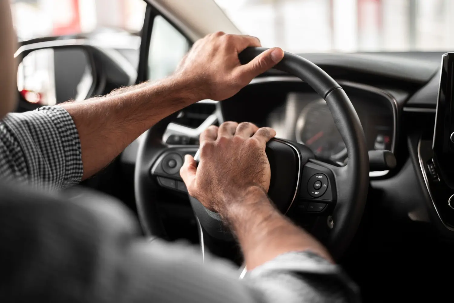 Foto aproximada de mãos de homem segurando um volante e pressionando a buzina, durante um dia ensolarado. O painel do carro é preto e o condutor usa uma camisa social quadriculada e dobrada até o cotovelo.