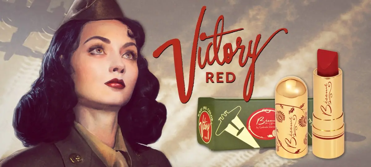 Uma imagem promocional de inspiração vintage para um batom chamado 'Victory Red' (Vermelho Vitória). À esquerda, o busto de uma mulher com cabelo escuro e ondulado, vestindo um uniforme militar estilo Segunda Guerra Mundial e um quepe, olhando para cima com uma expressão séria. Ela está usando um batom vermelho vibrante que combina com o tema. À direita, sobre um fundo militar em tons de sépia com silhuetas de aviões, aparece o texto estilizado 'Victory RED' em letras vermelhas e grandes. Abaixo do texto, há uma caixa verde-oliva com um desenho de um avião e dois tubos de batom dourados, um com a tampa e o outro sem, revelando o batom vermelho sólido. Os produtos pertencem à marca Bésame Cosmetics.