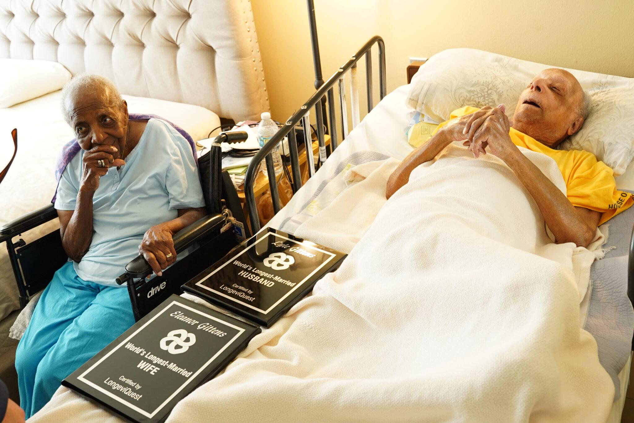 Imagem emocionada de idosos em um ambiente hospitalar, um deles na cama e outro em cadeira de rodas, celebrando bodas de casamento com placas comemorativas.