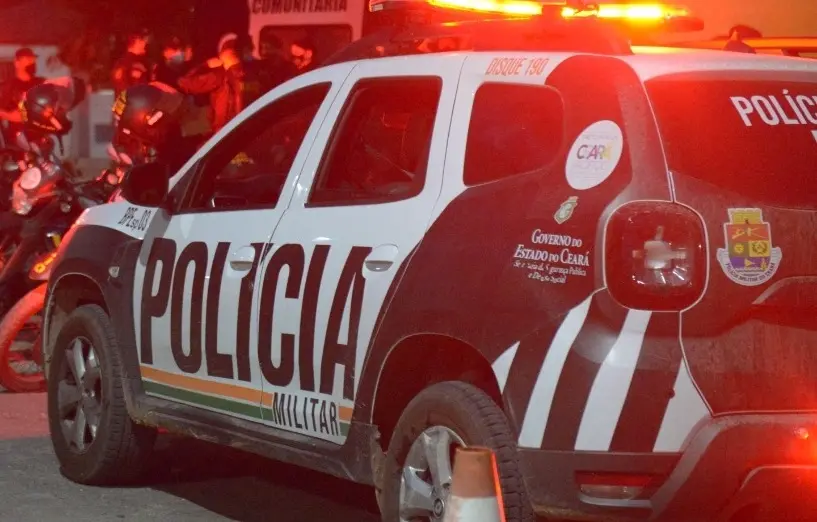 Foto de uma viatura da Polícia Militar do Ceará à noite. O veículo é branco e preto com adesivos do Governo do Ceará e é iluminado indiretamente por luz vermelha do intermitente. Ao fundo, há policiais com fardamento verde escuro.