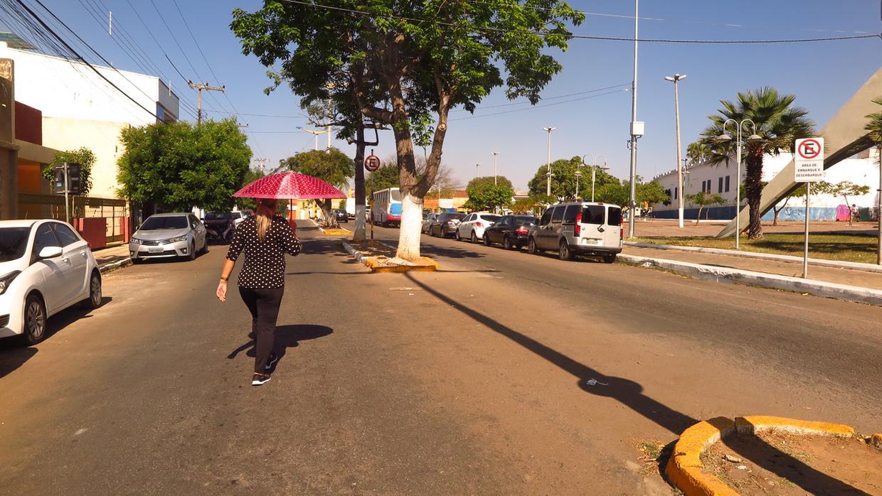 Fotografia em Juazeiro do Norte em dia de sol e calor, mostrando uma mulher caminhando pela rua com uma sombrinha vermelha para se proteger do sol.