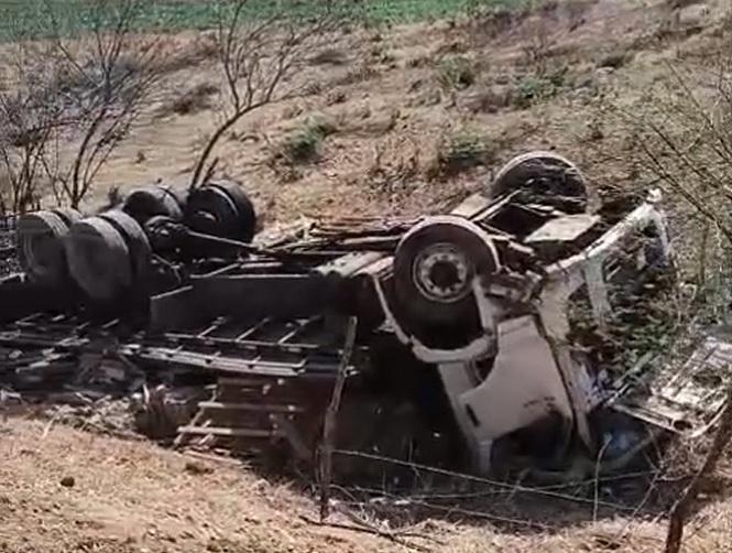 Imagem de um caminhão capotado na lateral de uma estrada de terra, em uma área de vegetação seca, resultado de um acidente.