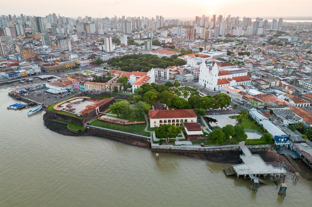 Na imagem, uma vista aérea da orla de Belém, Pará, Brasil, mostrando o Forte do Castelo e a Catedral Metropolitana de Belém (Sé) à beira de um grande rio. O centro histórico com telhados de terracota e edifícios coloniais se destaca contra a moderna paisagem urbana com arranha-céus ao fundo. A água do rio é de cor barrenta e o céu está em um tom alaranjado de pôr do sol ou amanhecer.
