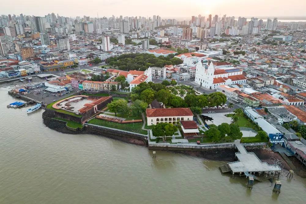 Na imagem, uma vista aérea da orla de Belém, Pará, Brasil, mostrando o Forte do Castelo e a Catedral Metropolitana de Belém (Sé) à beira de um grande rio. O centro histórico com telhados de terracota e edifícios coloniais se destaca contra a moderna paisagem urbana com arranha-céus ao fundo. A água do rio é de cor barrenta e o céu está em um tom alaranjado de pôr do sol ou amanhecer.