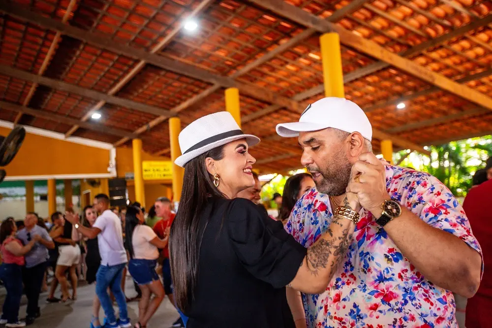 Na imagem, um casal sorridente dança próximo em um ambiente interno com teto de telhas. A mulher usa um chapéu fedora branco e brincos de argola, enquanto o homem veste um boné branco e uma camisa floral colorida. Ele tem tatuagens visíveis no braço. Outras pessoas estão dançando no fundo.