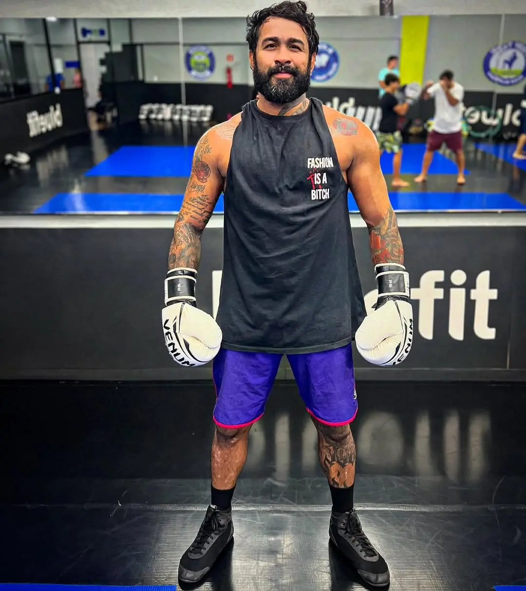 Imagem mostra Godofredo Pepey vestindo luvas de boxe e roupas de treino, posando em academia de boxe com sacos de pancada ao fundo.
