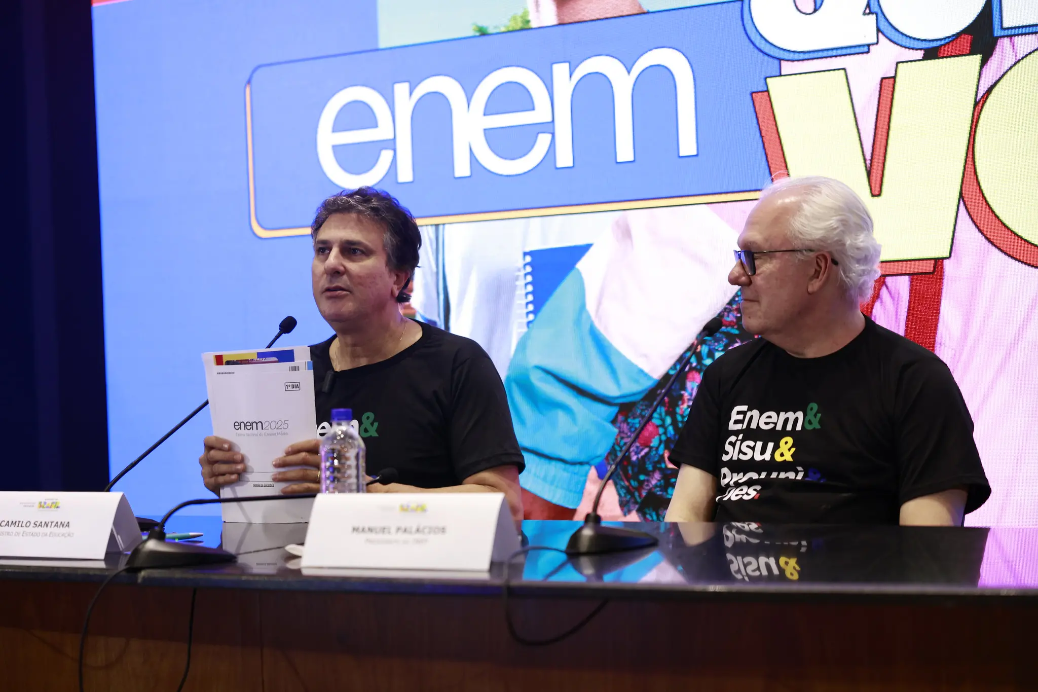 Uma foto de duas autoridades sentadas atrás de uma mesa em um evento. À esquerda está Camilo Santana, Ministro da Educação, segurando uma cópia do caderno de provas do Enem 2025. À direita está Manuel Palacios, ex-presidente do INEP. Ambos vestem camisetas pretas com logos relacionadas ao Enem/SiSU/ProUni. Um grande painel azul ao fundo exibe o logo 'enem'. O ambiente parece ser uma coletiva de imprensa ou balanço de evento.