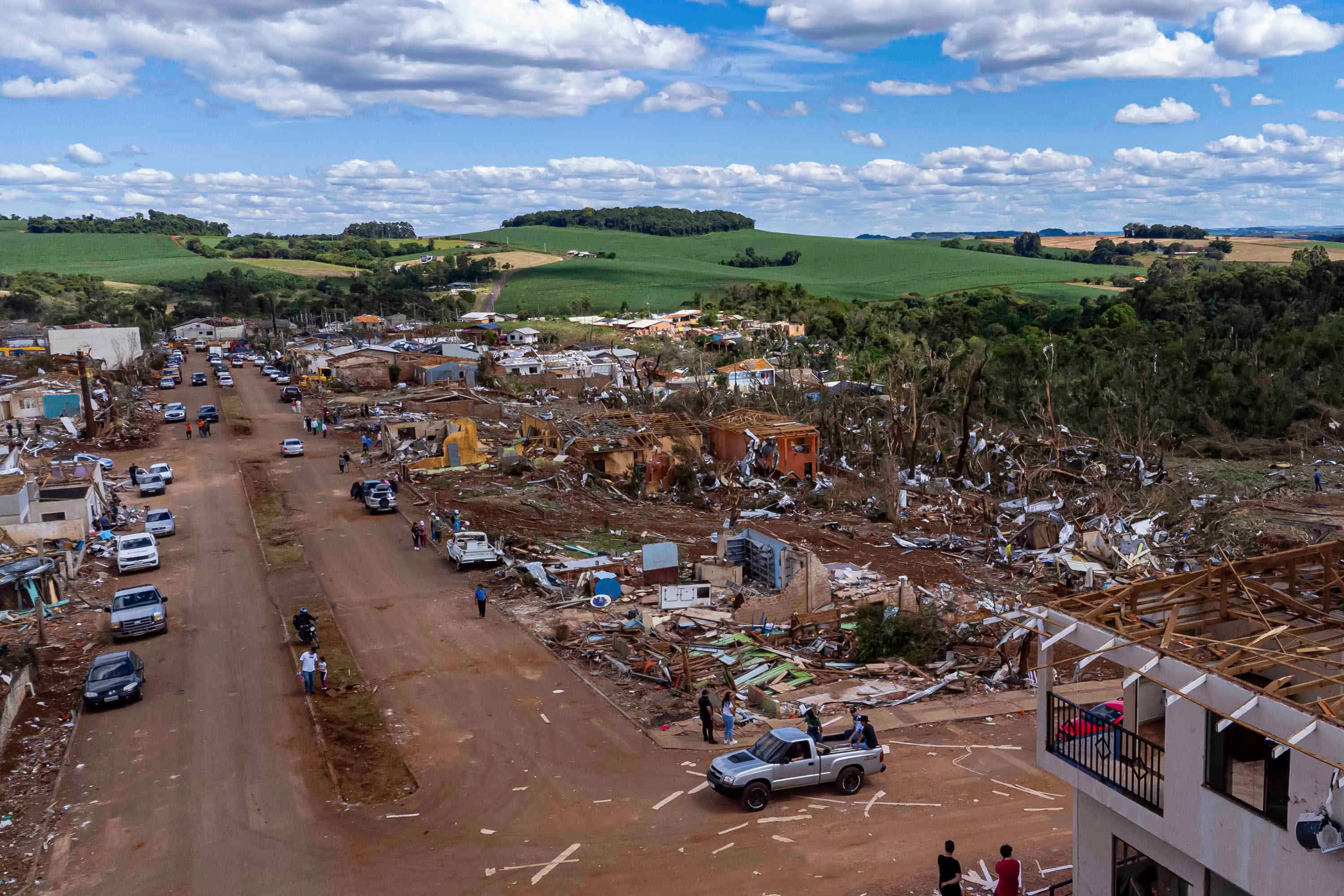 Vista aérea de uma área residencial em Rio Bonito do Iguaçu, Brasil, após ter sido devastada por um tornado. A imagem mostra uma rua de terra ladeada por destroços de casas completamente destruídas ou severamente danificadas, com telhados arrancados e paredes desmoronadas. Pessoas e veículos, incluindo várias caminhonetes, estão na rua e entre os escombros, sinalizando esforços de resgate ou limpeza. Ao fundo, áreas de vegetação e colinas verdes contrastam com a destruição na área urbana. O céu está parcialmente nublado, e a luz do sol sugere que a foto foi tirada durante o dia.