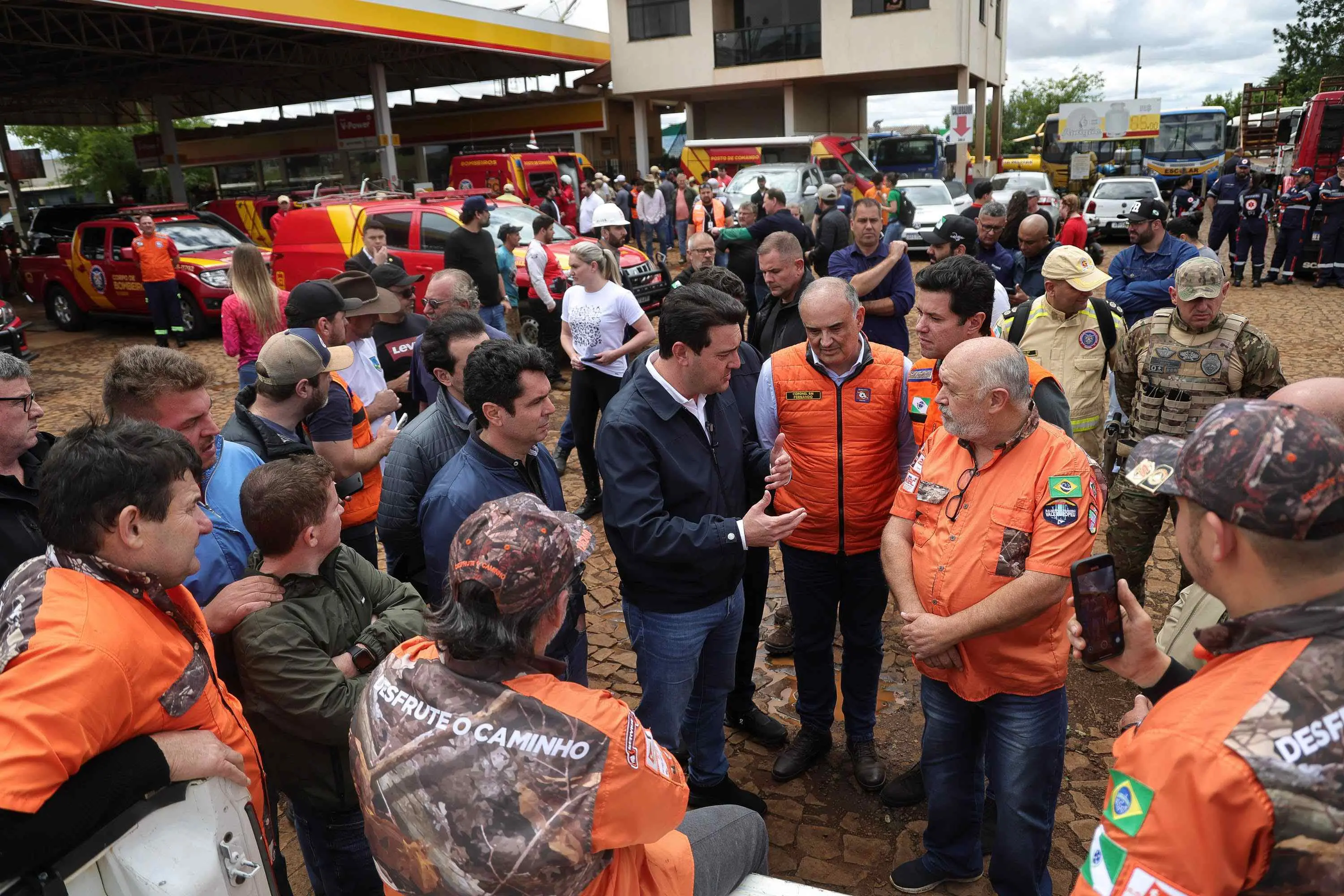 Grupo de pessoas, incluindo o governador do Rio Grande do Sul, Eduardo Leite, e equipes de resgate uniformizadas (principalmente em laranja), reunidas em um pátio. Eles estão em uma conversa intensa, com veículos de emergência ao fundo, sugerindo um briefing ou ponto de apoio durante uma situação de crise.