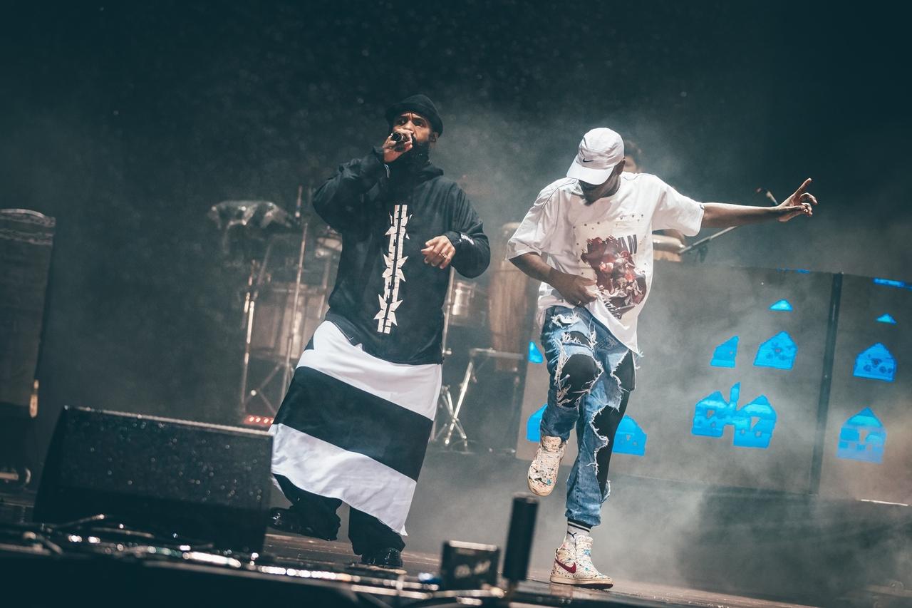 Foto de show noturno da banda BaianaSystem no Lollapalooza. Dois membros estão no palco, envoltos em fumaça de luzes de palco. À esquerda, um vocalista veste um gorro preto e uma longa túnica preta e branca listrada na horizontal, segurando um microfone enquanto canta. À direita, outro membro veste uma camiseta branca oversize, um boné branco e jeans rasgados, em uma pose dinâmica com um braço estendido. Ao fundo, vislumbram-se a bateria e instrumentos, com elementos de palco iluminados por uma luz azul.