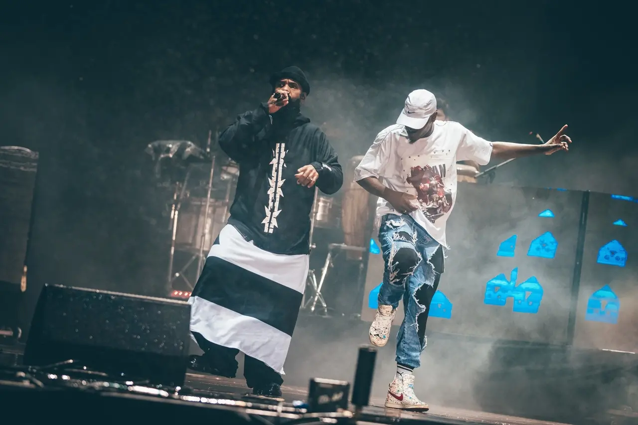 Foto de show noturno da banda BaianaSystem no Lollapalooza. Dois membros estão no palco, envoltos em fumaça de luzes de palco. À esquerda, um vocalista veste um gorro preto e uma longa túnica preta e branca listrada na horizontal, segurando um microfone enquanto canta. À direita, outro membro veste uma camiseta branca oversize, um boné branco e jeans rasgados, em uma pose dinâmica com um braço estendido. Ao fundo, vislumbram-se a bateria e instrumentos, com elementos de palco iluminados por uma luz azul.