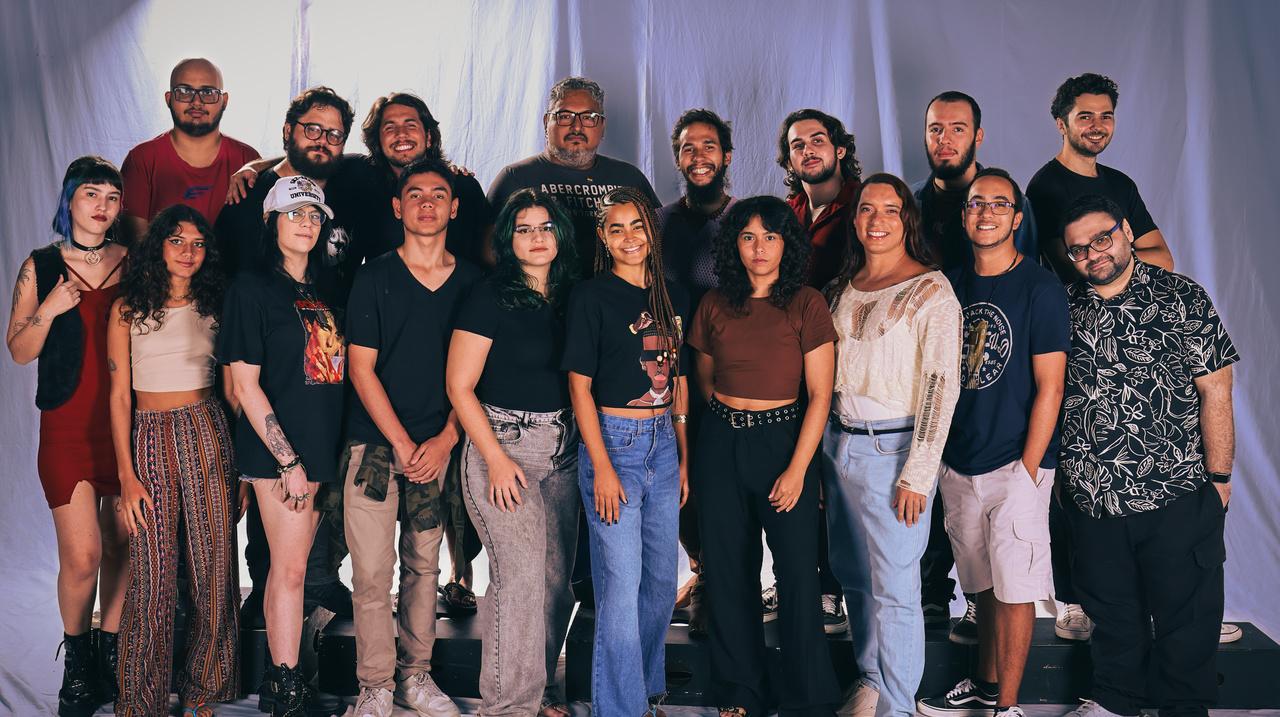 Uma foto de grupo de 18 cineastas, jovens adultos e de adultos, posando em frente a um fundo de cor neutra com cortinas drapeadas. A maioria está sorrindo. O grupo inclui pessoas de diversas etnias e tipos de corpo, vestidas com roupas casuais, variando de camisetas e calças largas a tops e saias/calças estampadas.