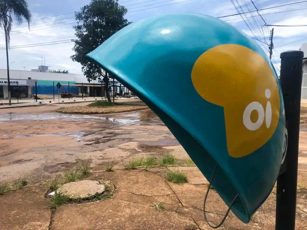 Orelhão azul e amarelo (telefone público) da operadora Oi, em um ambiente externo com chão de terra e cimento rachado. Ao fundo, há um prédio municipal parcialmente visível sob um céu claro.