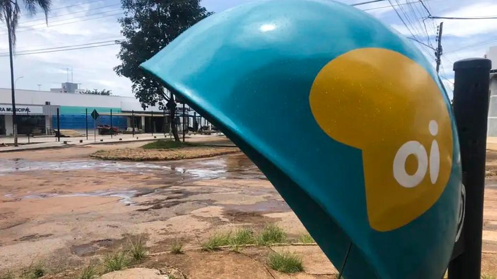 Orelhão azul e amarelo (telefone público) da operadora Oi, em um ambiente externo com chão de terra e cimento rachado. Ao fundo, há um prédio municipal parcialmente visível sob um céu claro.