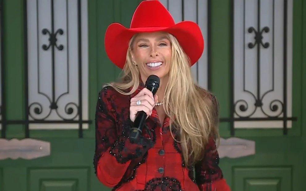 Foto de Adriane Galisteu de roupa vermelha e sorrindo com um microfone na mão apresentando A Fazenda.