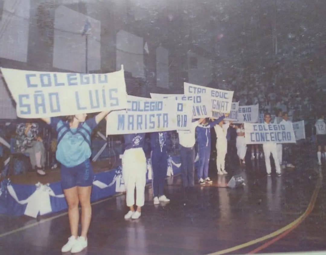Imagem colorida e antiga, com baixa luminosidade, em um ginásio de esportes. Pessoas em primeiro plano seguram faixas brancas com nomes de colégios em azul, como COLÉGIO SÃO LUÍS e COLÉGIO MARISTA. O público lota as arquibancadas ao fundo, sugerindo um evento esportivo ou cultural.
