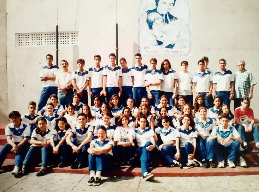 Foto antiga, datada de 1996, com um grande grupo de estudantes e alguns adultos posando juntos. A maioria dos estudantes veste o mesmo uniforme branco e azul. O grupo está sentado e em pé em degraus. Ao fundo, uma parede clara com um mural de fundo azul.