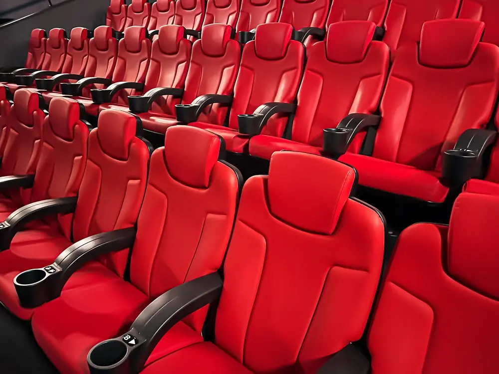 Na imagem, fileiras de poltronas de cinema vazias e luxuosas, estofadas em couro vermelho brilhante. As cadeiras têm apoios de cabeça altos e apoios de braço pretos com porta-copos embutidos. A perspectiva é em ângulo, focando nas fileiras mais próximas, dando a sensação de um cinema moderno ou sala de projeção VIP.